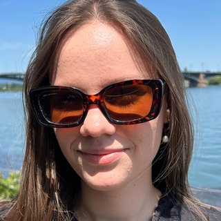 Junge Frau mit Sonnenbrille steht vor Wasser.