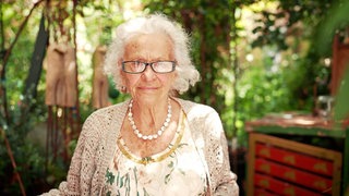 Alte Frau mit weißen Haaren und Brille in einem Garten.