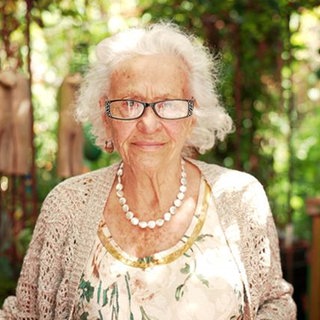 Alte Frau mit weißen Haaren und Brille in einem Garten.