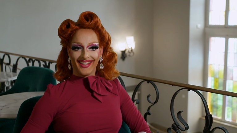 Dragqueen mit roten Haaren und einem bordeauxroten Kleid lächelt in die Kamera.