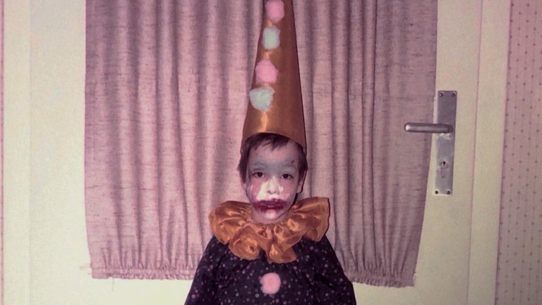 Ein kleines Kind, verkleidet im Clowns Kostüm lächelt in die Kamera. Bei dem Kind auf dem Bild handelt es sich um Chardonnay.