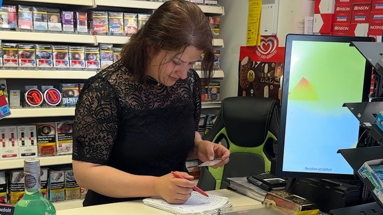 Eine Frau mit braunen Haaren und schwarzem Oberteil steht hinter einer Ladentheke und schreibt.