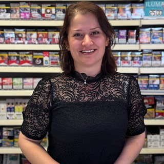 Eine Frau mit braunen Haaren und schwarzem Oberteil lächelt in Kamera. Hinter ihr sind Regale mit Waren.