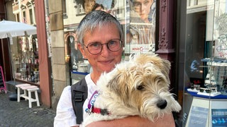 Eine Frau mit kurzen grauen Haaren und Brille hält grinsend einen Hund auf dem Arm. Heide aus Grebenau war früher lange sehr einsam, ist aber jetzt seit 20 Jahren glücklich alleinstehend.