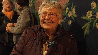 Eine Seniorin mit Brille und kurzen, grauen Haaren lächelt freundlich in die Kamera.