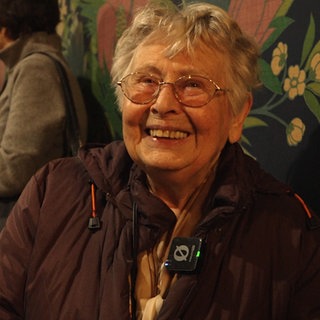 Eine Seniorin mit Brille und kurzen, grauen Haaren lächelt freundlich in die Kamera.