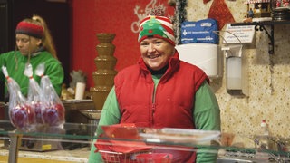 Eine Frau steht in ihrem Verkaufstand auf dem Weihnachtsmarkt. Sie trägt eine Weste, ein Stirnband und lächelt in die Kamera.