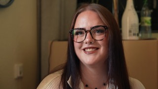 Eine junge Frau mit glatten Haaren und Brille sitzt in einem Raum und lächelt in die Kamera.