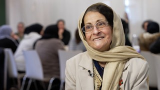 Lächelnde ältere Dame mit Kopftuch. Im Hintergrund sitzen Menschen um einen Tisch