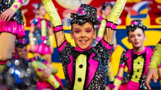 Junges Mädchen in buntem Outfit, glitzerndem Hut und buntem Make-Up lächelt in Kamera.