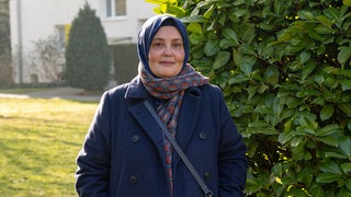 Frau mit blauem Kopftuch, buntem Schal und blauem Mantel schaut in Kamera.