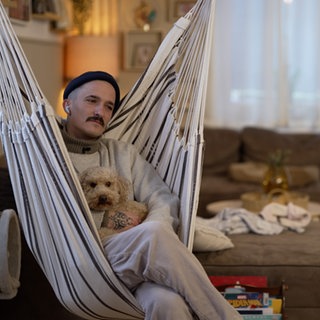 Junger Mann mit Hund im Arm im Wohnzimmer in einer Hängematte