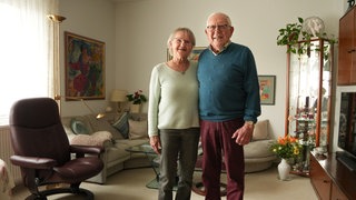 Frau und Mann mit weißen Haaren stehen in ihrem Wohnzimmer.