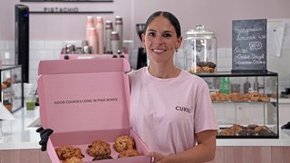 Junge Frau mit braunen Haaren steht vor einer Ladentheke. In der Hand hält sie einen großen Karton mit Cookies.