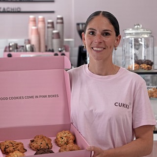 Junge Frau mit braunen Haaren steht vor einer Ladentheke. In der Hand hält sie einen großen Karton mit Cookies.