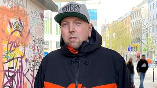 Simon steht neben dem Stand vom „Paule Club” in Stuttgart unter der Paulinenbrücke. Er ist Sprecher und Mitgründer des „Paule Clubs”. Er trägt eine graue Cap und eine schwarz-orangefarbene Jacke. Er schaut in die Kamera. 