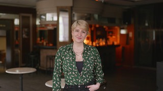 Anna aus spricht über ihre Bi+sexualität. Sie steht in einer Bar, im Hintergrund ist die Theke zu sehen. Sie trägt eine grüne Bluse und hat eine Hand in ihre Hosentaschen gesteckt. Sie lächelt in die Kamera.