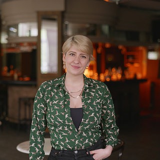 Anna aus spricht über ihre Bi+sexualität. Sie steht in einer Bar, im Hintergrund ist die Theke zu sehen. Sie trägt eine grüne Bluse und hat eine Hand in ihre Hosentaschen gesteckt. Sie lächelt in die Kamera.
