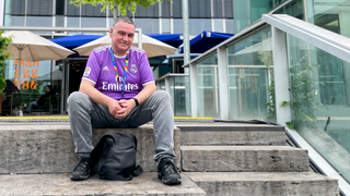 Wir treffen Gerhard beim Public Viewing auf dem Stuttgarter Schlossplatz. Er trägt ein lila Fußballtrikot. Er sitzt auf einer Treppe und lächelt in die Kamera.