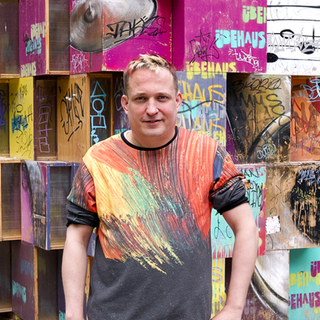 Jean-Marc ist Musiker. Er hat Tourette und räumt mit Vorurteilen auf. Er steht vor einer Wand, die aus bunten Würfeln besteht. Er schaut in die Kamera. 