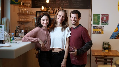 Clara, Imogen und Jadu leben polyamor. Sie stehen in ihrem Restaurant und lächeln in die Kamera.