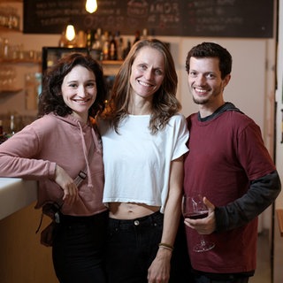 Clara, Imogen und Jadu leben polyamor. Sie stehen in ihrem Restaurant und lächeln in die Kamera.