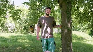  Jerome arbeitet als Gärtner. Er lehnt sich an einen Baum und schaut in die Kamera. 