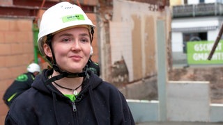 Martha ist 18 Jahre alt und macht nach ihrem Abitur eine Ausbildung zur Stahlbetonbauerin. Sie steht im Vordergrund und schaut in die Kamera. Sie trägt einen Helm und im Hintergrund sind ihre Kollegen zu sehen.