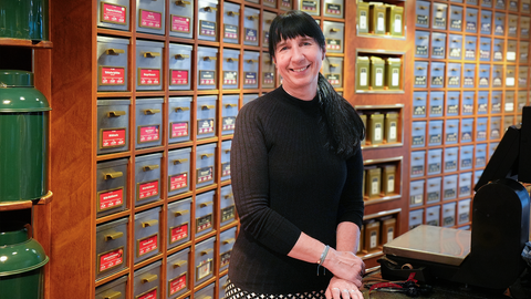 Margit steht hinter der Kasse in ihrem Tee-Fachgeschäft. Im Hintergrund stehen große Ladenregale gefüllt mit viel Tee. Sie lacht in die Kamera.