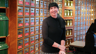 Margit steht hinter der Kasse in ihrem Tee-Fachgeschäft. Im Hintergrund stehen große Ladenregale gefüllt mit viel Tee. Sie lacht in die Kamera.