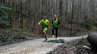 Der blinde Läufer Pierre und sein Guide beim Joggen im Wald. Sie springen über einen Ast und sind durch eine Schlaufe an ihren Händen miteinander verbunden.
