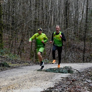 Der blinde Läufer Pierre und sein Guide beim Joggen im Wald. Sie springen über einen Ast und sind durch eine Schlaufe an ihren Händen miteinander verbunden.