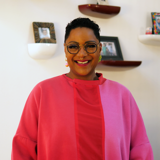 Jennifer, eine Schwarze Frau Mitte 40, strahlt in die Kamera. Sie trägt ein pink-rotes, leuchtendes Oberteil, hinter ihr an der Wand hängen Familienfotos.