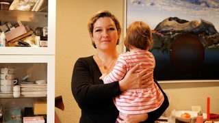 Die alleinerziehende Mama Hanna, hat ihre kleine Tochter auf dem Arm. Sie steht in ihrem Esszimmer und schaut in die Kamera.