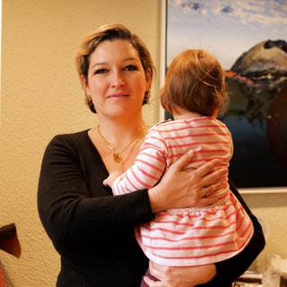 Die alleinerziehende Mama Hanna, hat ihre kleine Tochter auf dem Arm. Sie steht in ihrem Esszimmer und schaut in die Kamera.