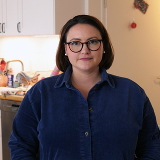 Die alleinerziehende Mutter Laura schaut mit ernstem Blick in die Kamera. Sie trägt glatte dunkle Haare bis zu den Schultern und eine Brille mit schwarzem Gestell. Hinter ihr ist ihre helle, ordentliche Wohnung zu sehen.