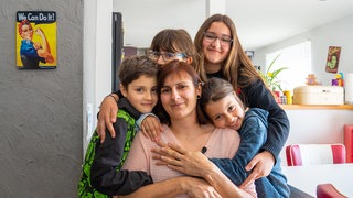 Frau in der Mitte des Bildes wird von vier Kindern von hinten umarmt.