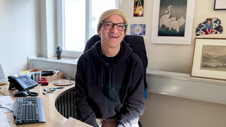 Wolfgang ist 55, mit einer dunklen Kapuzenjacke und einer bunten Wollmütze sitzt er auf seinem Bürostuhl am Schreibtisch