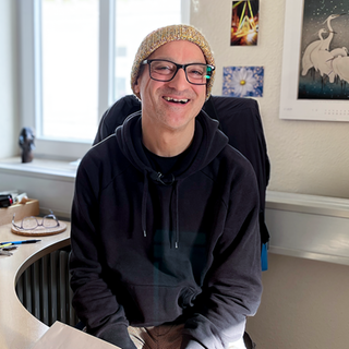 Wolfgang ist 55, mit einer dunklen Kapuzenjacke und einer bunten Wollmütze sitzt er auf seinem Bürostuhl am Schreibtisch