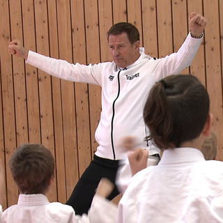 Eugen trägt eine weiße Sportjacke, auf der ‚Trainer‘ steht und macht Kindern in seiner Kampfkunstschule eine Ju-Jitsu-Übung vor.