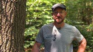 Sven in einem T-Shirt mit der Aufschrift „Schwäbischer Waldschrat“, er steht im Wald und schaut direkt in die Kamera.
