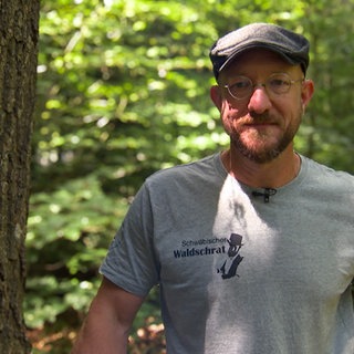 Sven in einem T-Shirt mit der Aufschrift „Schwäbischer Waldschrat“, er steht im Wald und schaut direkt in die Kamera.