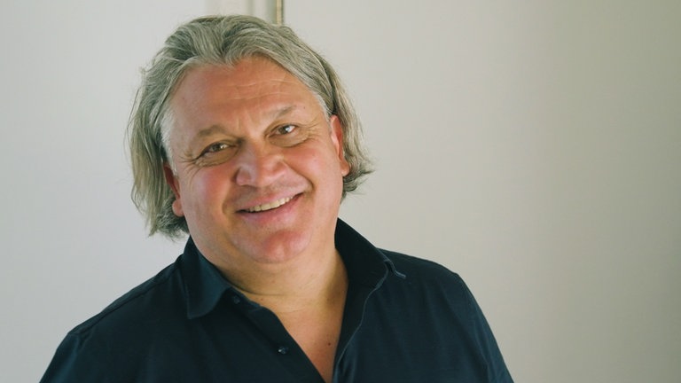 Mann mit längerem grauen Haar steht vor weißer Wand und lächelt in die Kamera.