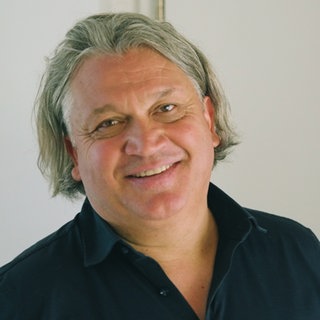 Mann mit längerem grauen Haar steht vor weißer Wand und lächelt in die Kamera.