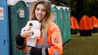 Diana trägt eine orange-graue Arbeitskleidung und hält mehrere Rollen Toilettenpapier in den Armen. Sie lächelt in die Kamera. Im Hintergrund sind bunte mobile Toilettenkabinen zu sehen - blaue, grüne und orangefarbene. 
