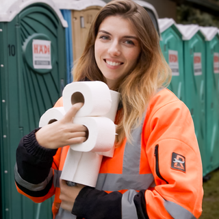 Diana trägt eine orange-graue Arbeitskleidung und hält mehrere Rollen Toilettenpapier in den Armen. Sie lächelt in die Kamera. Im Hintergrund sind bunte mobile Toilettenkabinen zu sehen - blaue, grüne und orangefarbene. 