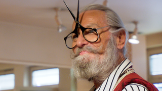 Der Figaro Uli steht vor seinem Friseursalon in Esslingen und lacht in die Kamera. Er trägt lange Gewänder in Weiß und Rot und hält einen roten Fächer in der Hand.  