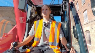Kranführerin Annika sitzt auf einer Baustelle in ihrem Kran. 