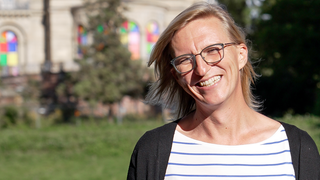Tanja steht in einem Park, hinter ihr eine bunte Villa, sie trägt eine Brille und ein blau-weiß gestreiftes T-Shirt, sie lächelt übers ganze Gesicht. Heute als Frau fühlt sie sich wohl in ihrem Körper. Doch als ihr Vater noch lebte, hat sie sich nicht getraut, sie selbst zu sein. 