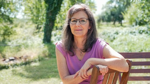 Tanja trägt eine Brille und lächelt schwach in die Kamera. Sie sitzt an einem Sommertag im Grünen auf einem Stuhl und spricht über den Tod ihres Sohnes und ihre Trauer.